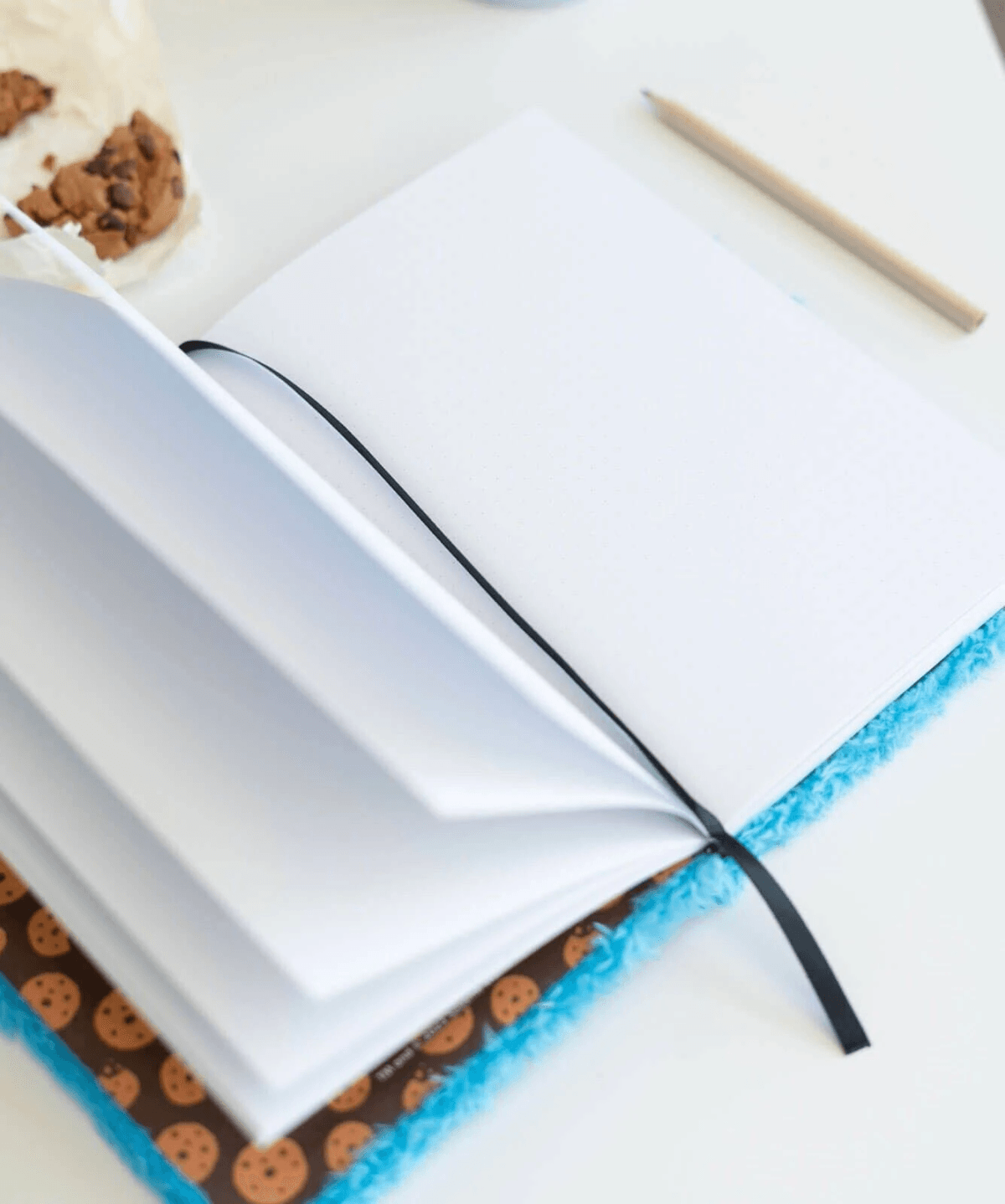 Un cuaderno abierto con borde azul peludo y estampado de galletas, junto a un lápiz y una galleta.