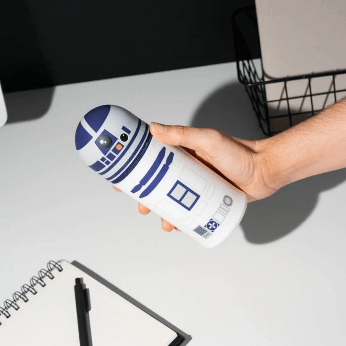 Una mano sostiene un recipiente blanco con forma de R2-D2 y acentos azules sobre un escritorio.