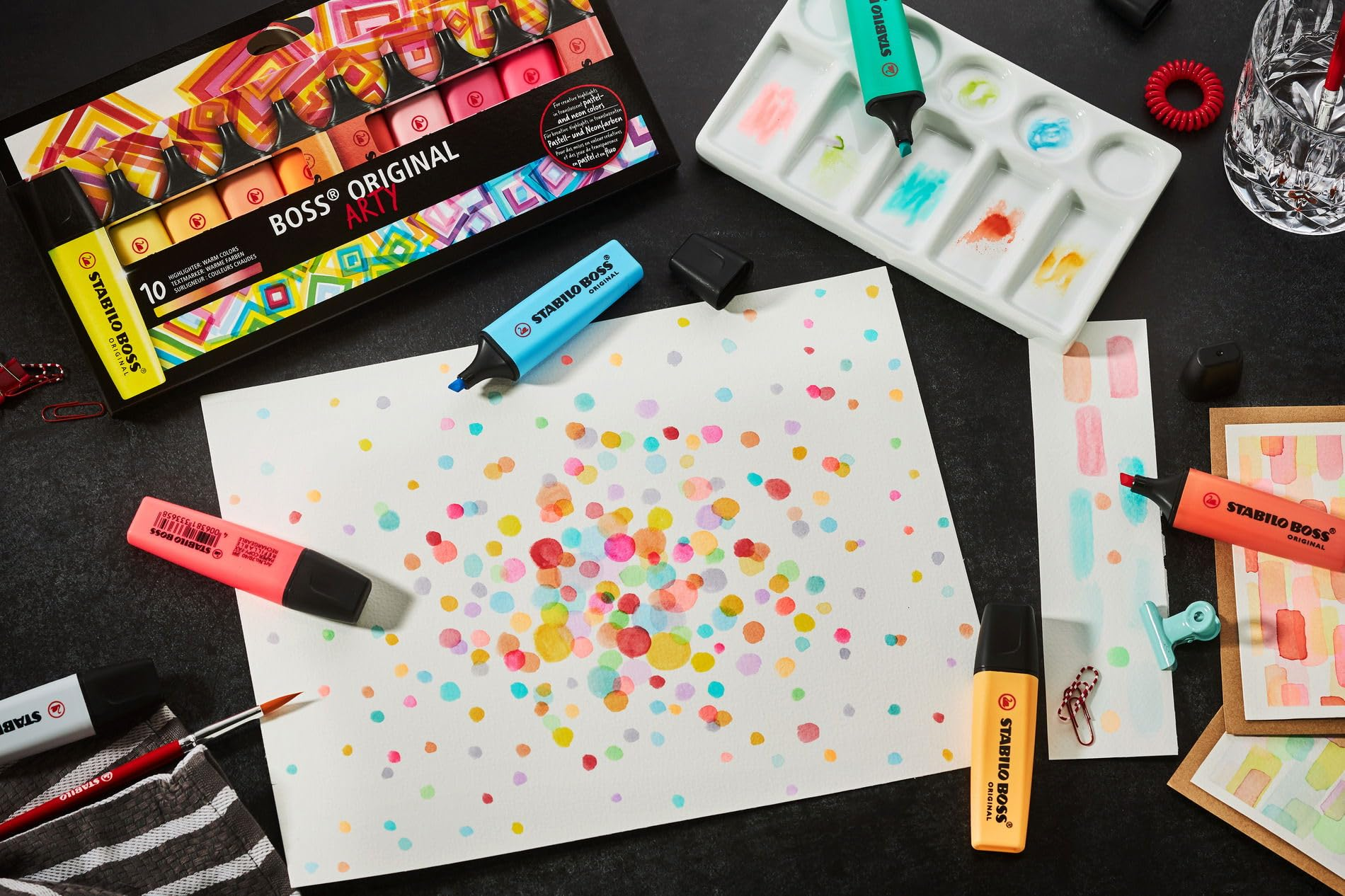 Bunte Punkte auf Papier, Stifte, eine Palette und ein Glas Wasser.