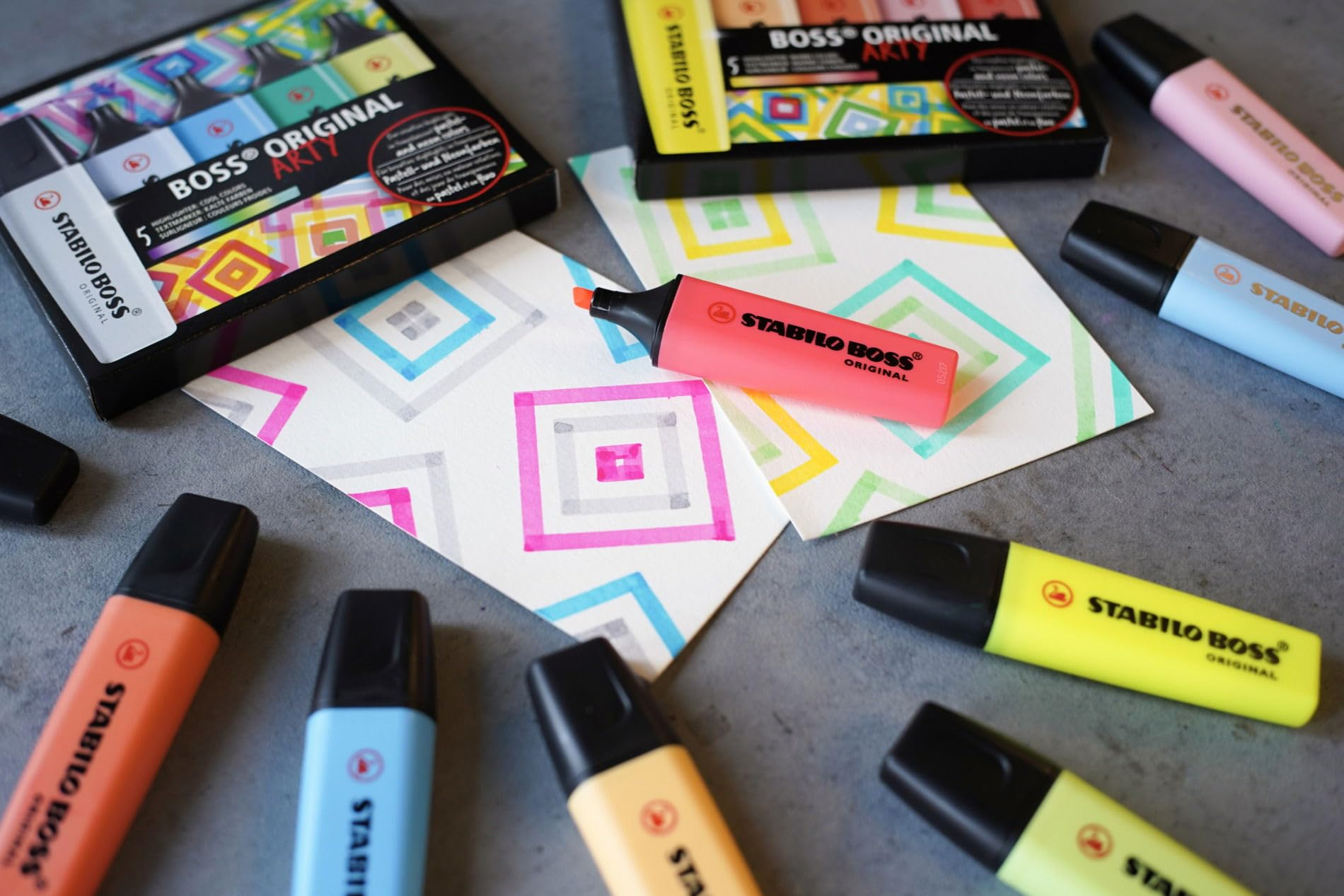 Bunte Stabilo Boss Textmarker liegen auf einer grauen Oberfläche mit gemusterten Papieren.