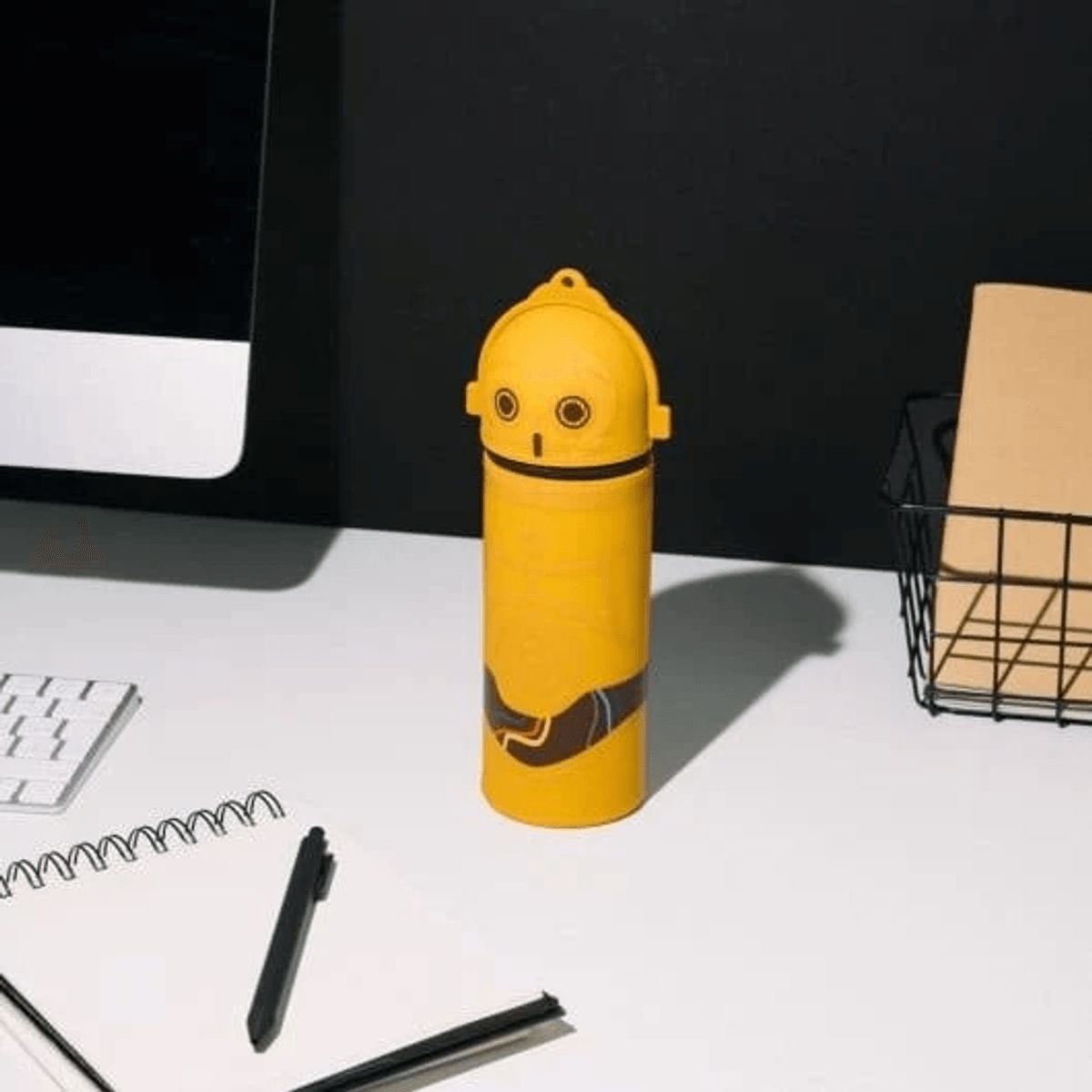 Portavasos amarillo estilo C-3PO sobre un escritorio con teclado y cuaderno.