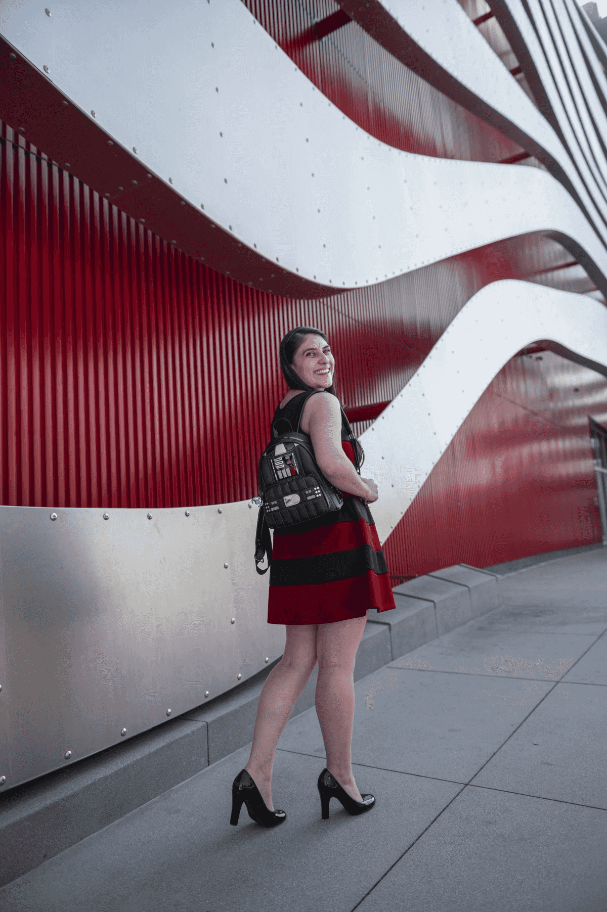 Una mujer con vestido a rayas rojas y negras sonríe, lleva mochila, frente a edificio rojo y plateado.