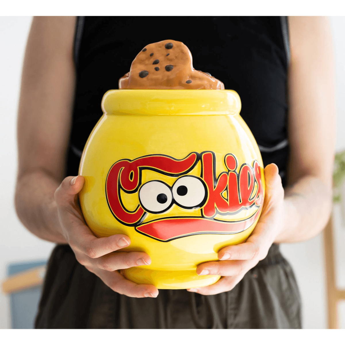 Tarro de galletas amarillo con ojos de dibujos animados y texto rojo "Cookies" sostenido por manos.