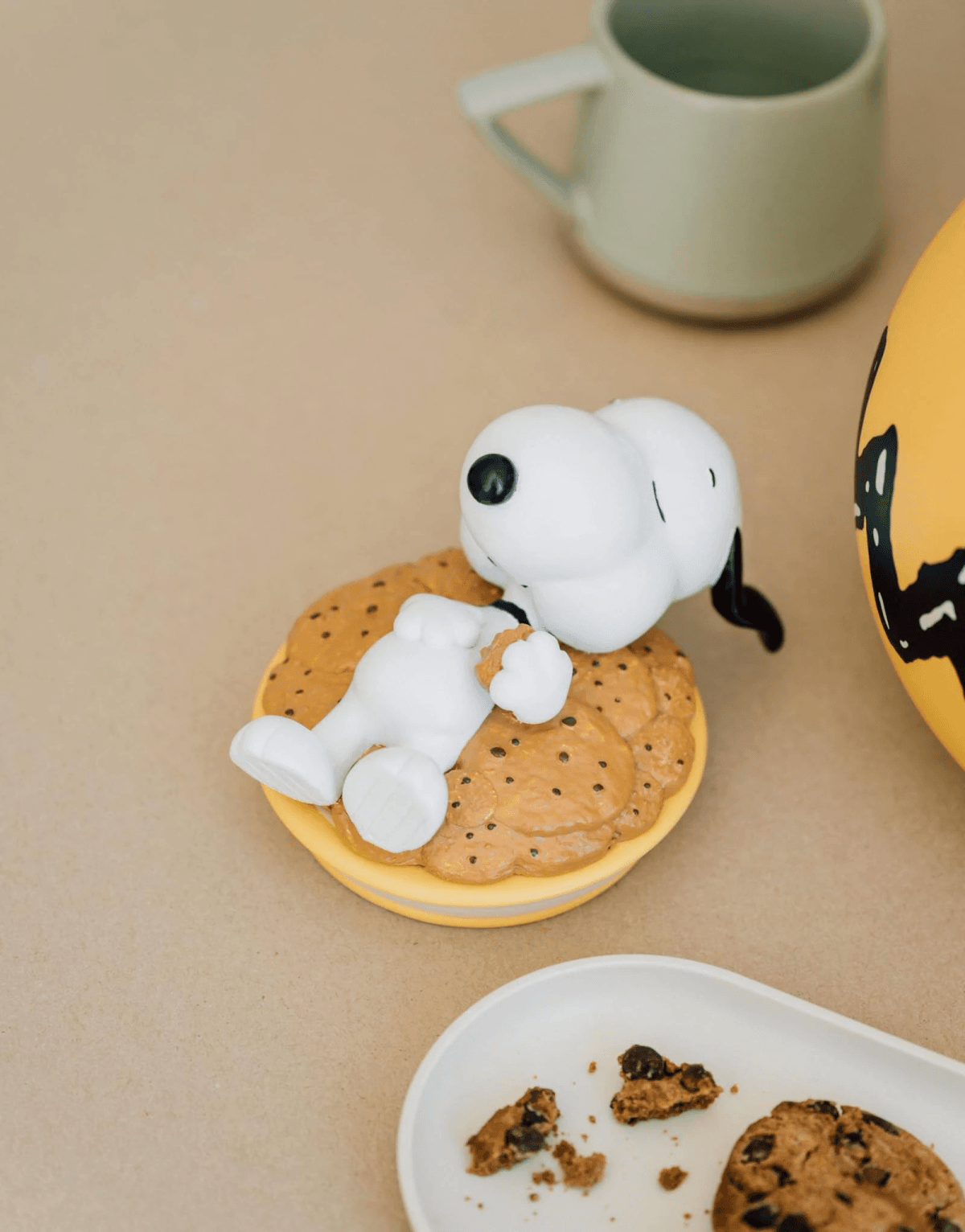 Una figura de Snoopy yace sobre una galleta, cerca de una taza y un objeto amarillo.