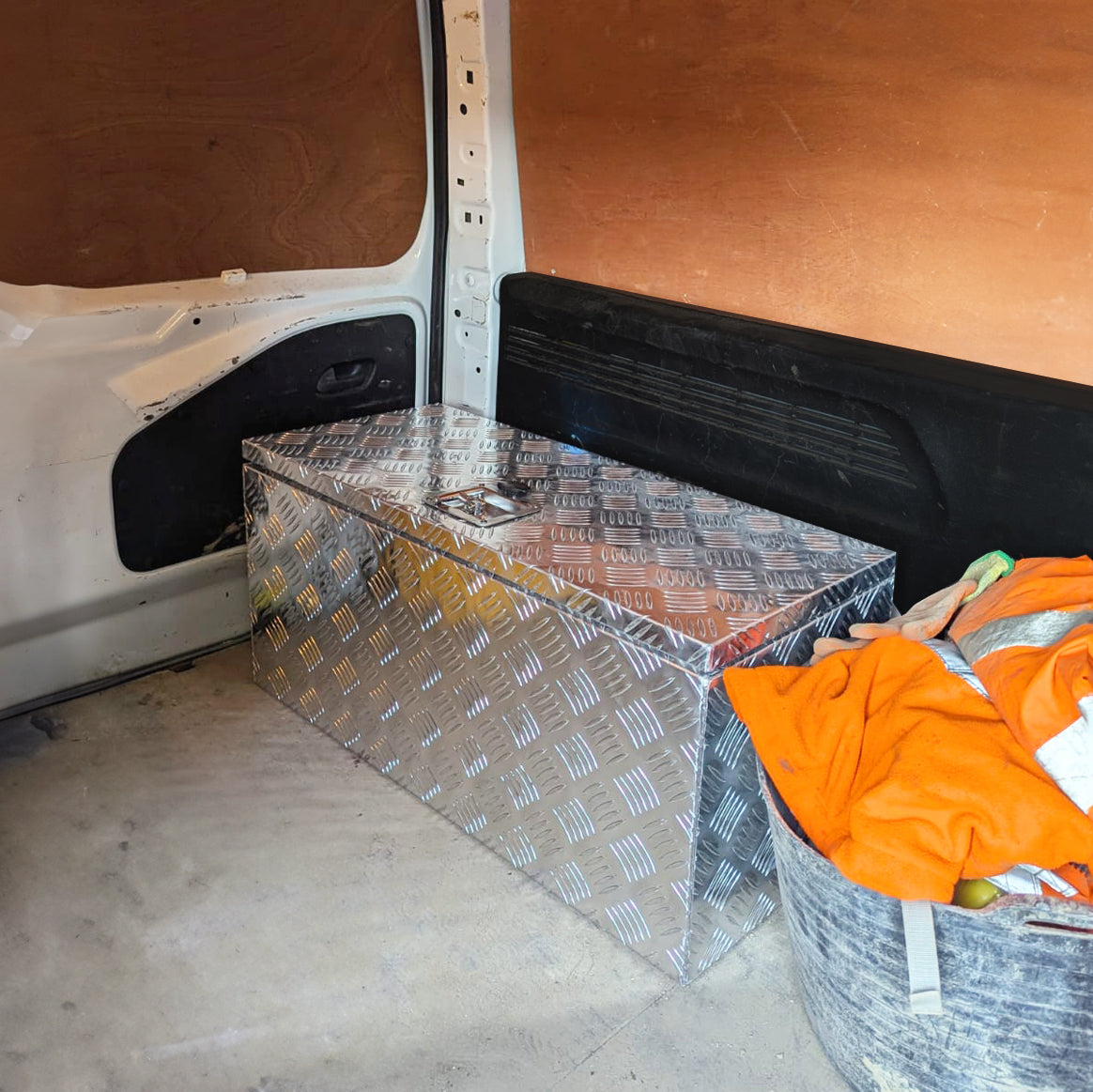 Una caja de herramientas plateada está en el área de carga de una furgoneta blanca, junto a un contenedor con ropa naranja.