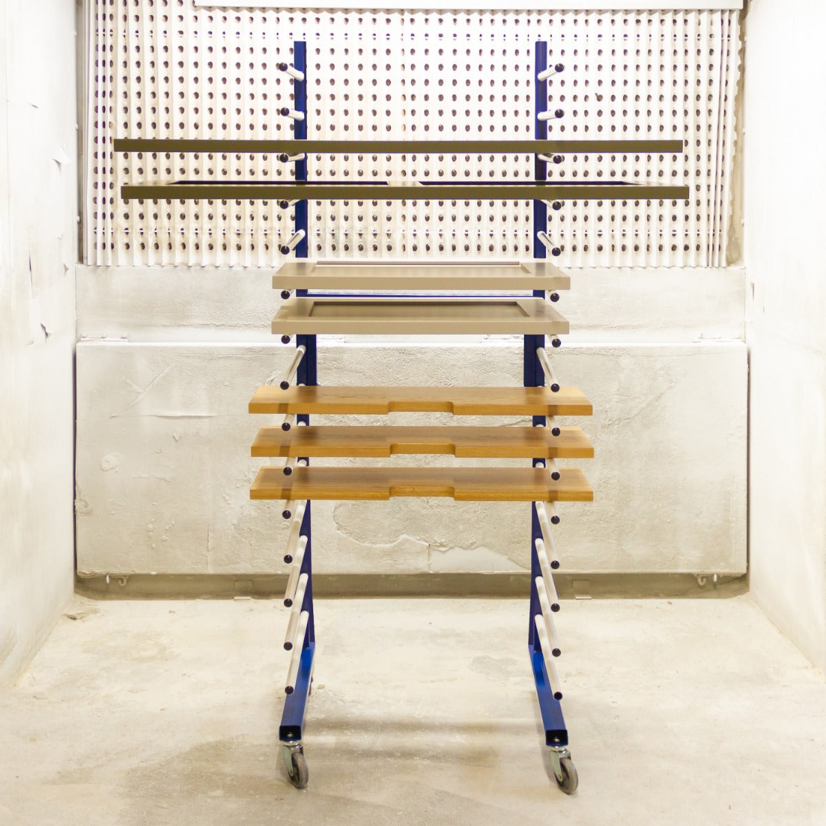Un carrito azul con estantes de madera y barras metálicas frente a un panel perforado.