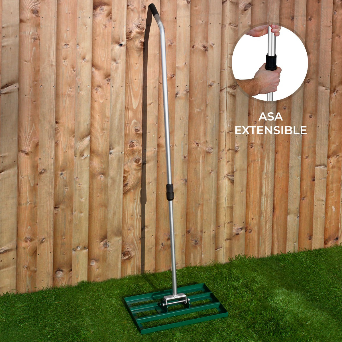 Un aireador de césped verde con mango extensible de aluminio sobre césped frente a valla de madera.