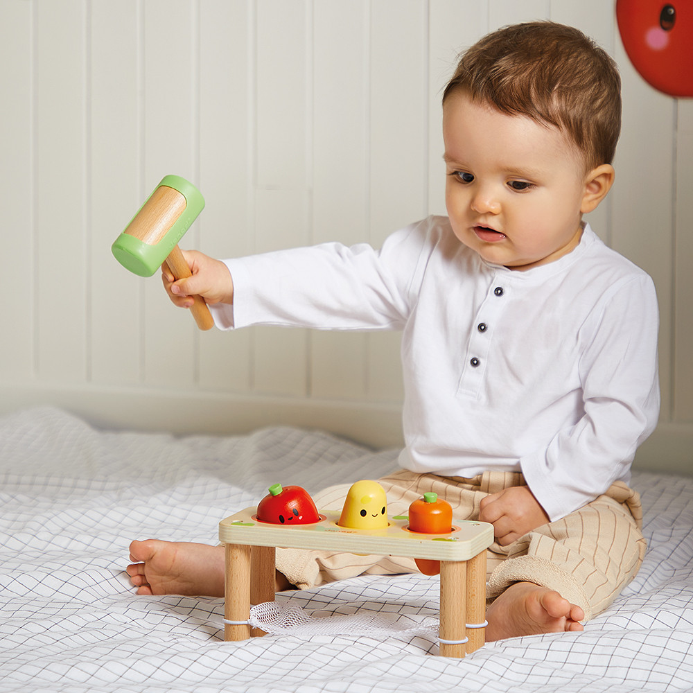Ein Baby spielt mit einem Holzhammer und einem Spielzeugtisch mit Früchten.
