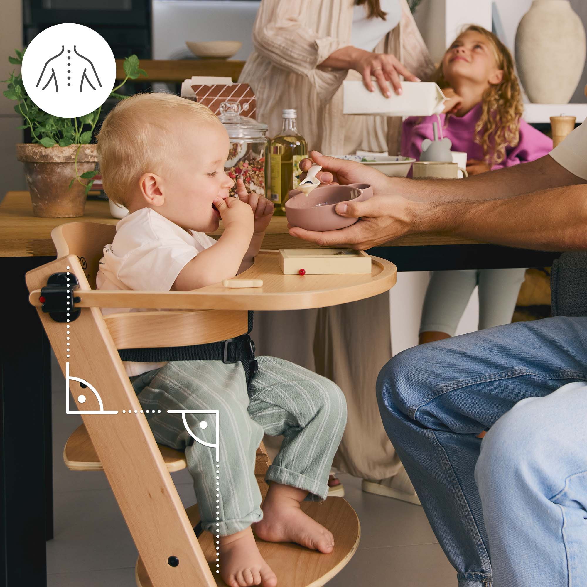 Ein Baby sitzt in einem Hochstuhl, während ein Erwachsener es füttert. Eine Familie ist im Hintergrund zu sehen.