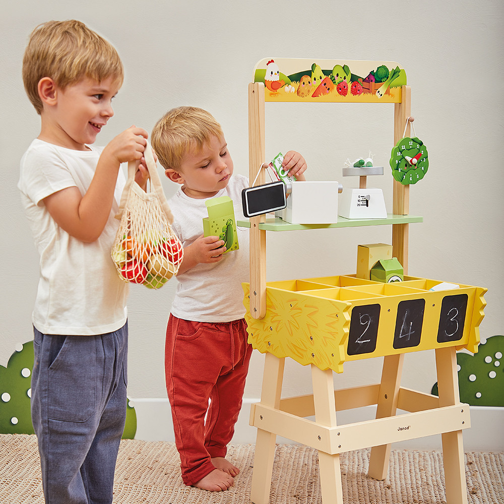 Zwei Jungen spielen an einem Holz-Kaufladen mit Spielzeuggemüse und einer Waage.