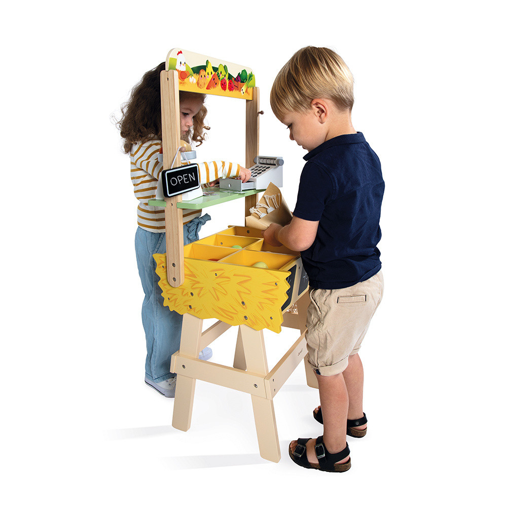 Zwei Kinder spielen an einem Spielzeugmarktstand. Ein Mädchen bedient, ein Junge packt Waren ein.
