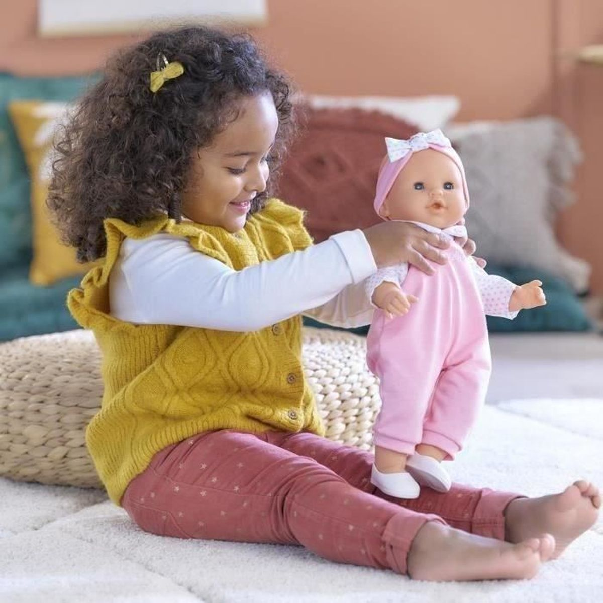 Ein junges Mädchen spielt mit einer Puppe in rosa Latzhose und Schleife, sitzend auf einem Teppich.