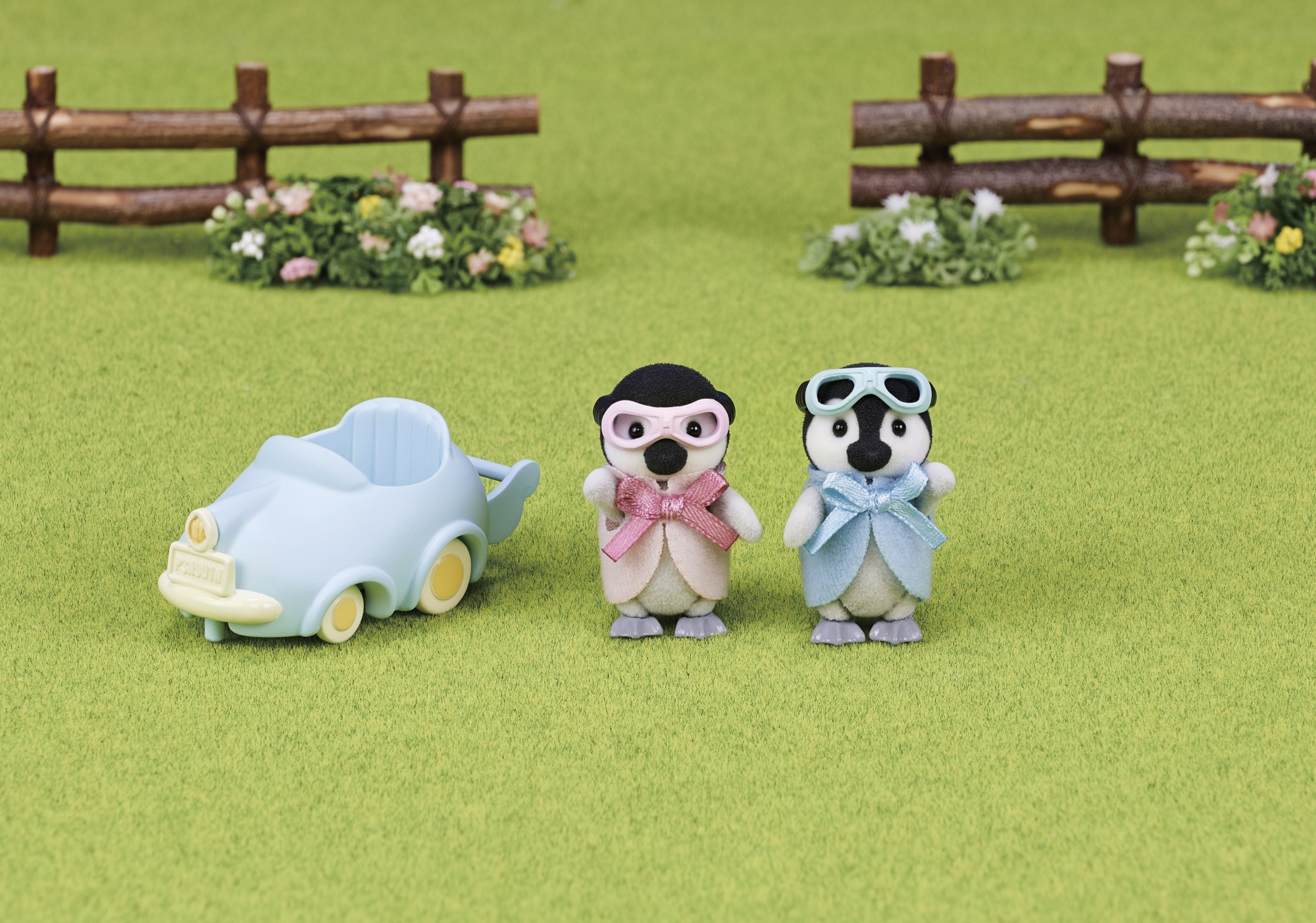 Dos figuras de pingüinos con ropa y gafas junto a un coche de juguete azul sobre hierba verde.