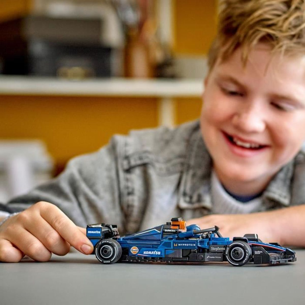 Bir çocuk masada mavi bir Lego Formula 1 arabasıyla oynuyor.