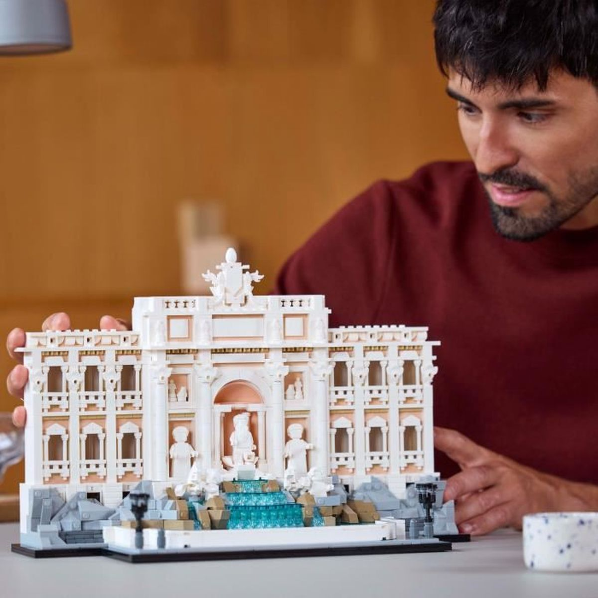 Bir adam mavi su öğeleriyle detaylı bir Lego Trevi Çeşmesi modeli tutuyor.