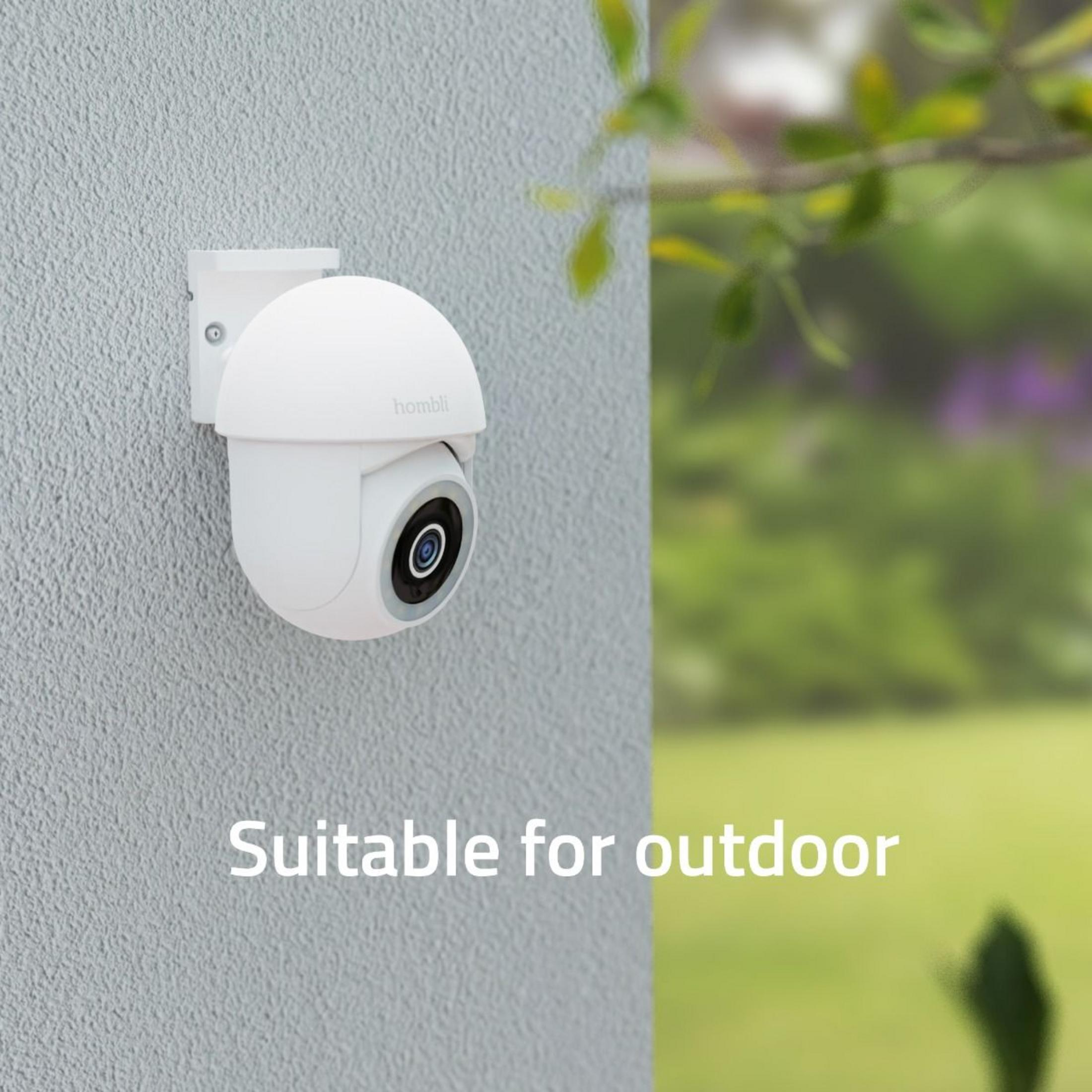 Eine weiße Hombli-Kamera ist an einer Wand montiert. Draußen ist ein Garten zu sehen.