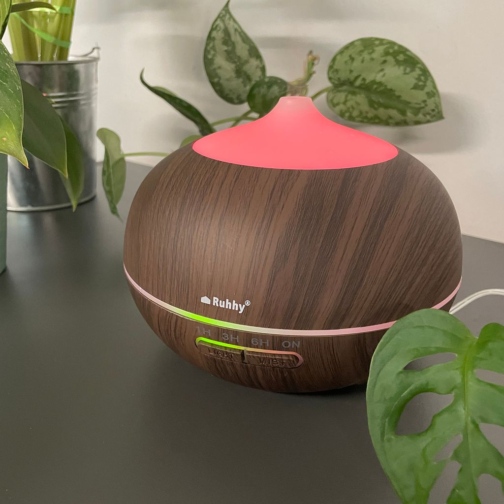 Ein brauner Diffusor mit Holzmaserung leuchtet pink und grün, mit Pflanzen im Hintergrund.