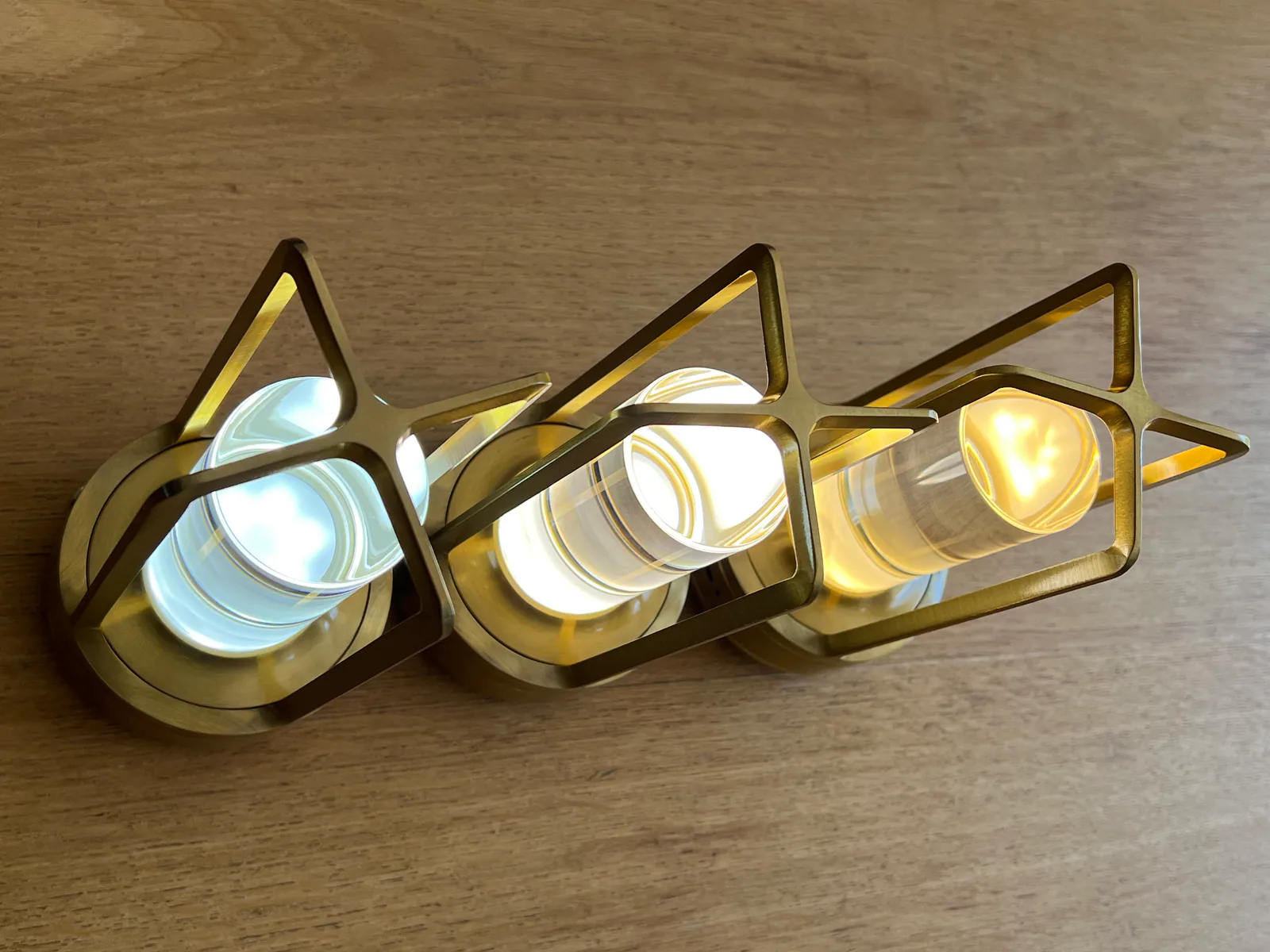 Eine moderne Messing-Wandleuchte mit drei Glaszylindern und Glühbirnen auf Holz.