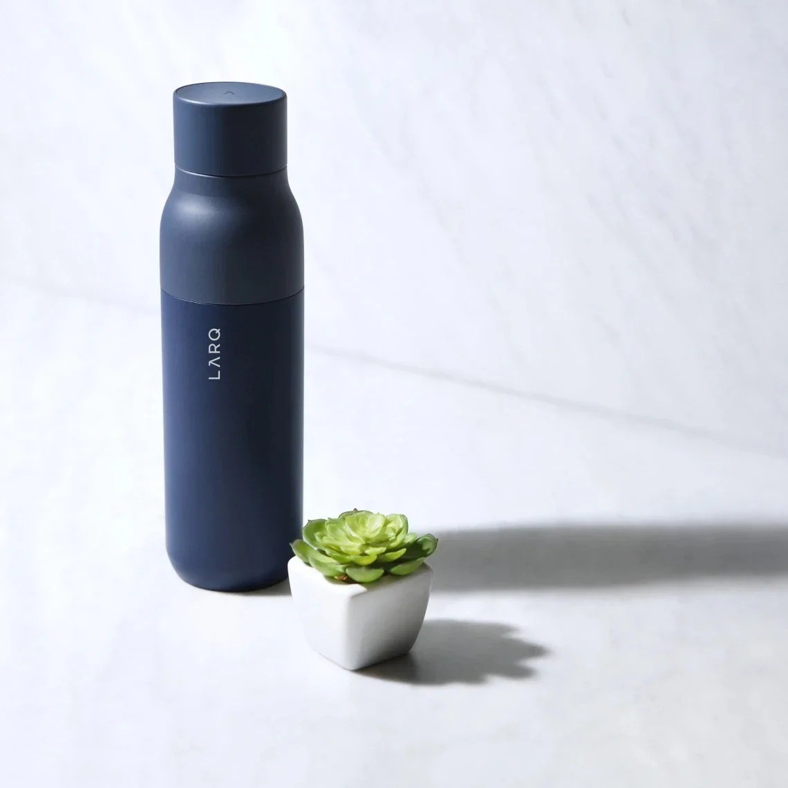 Dunkelblaue LARQ-Flasche neben einer kleinen Sukkulente in einem weißen Topf auf Marmor.