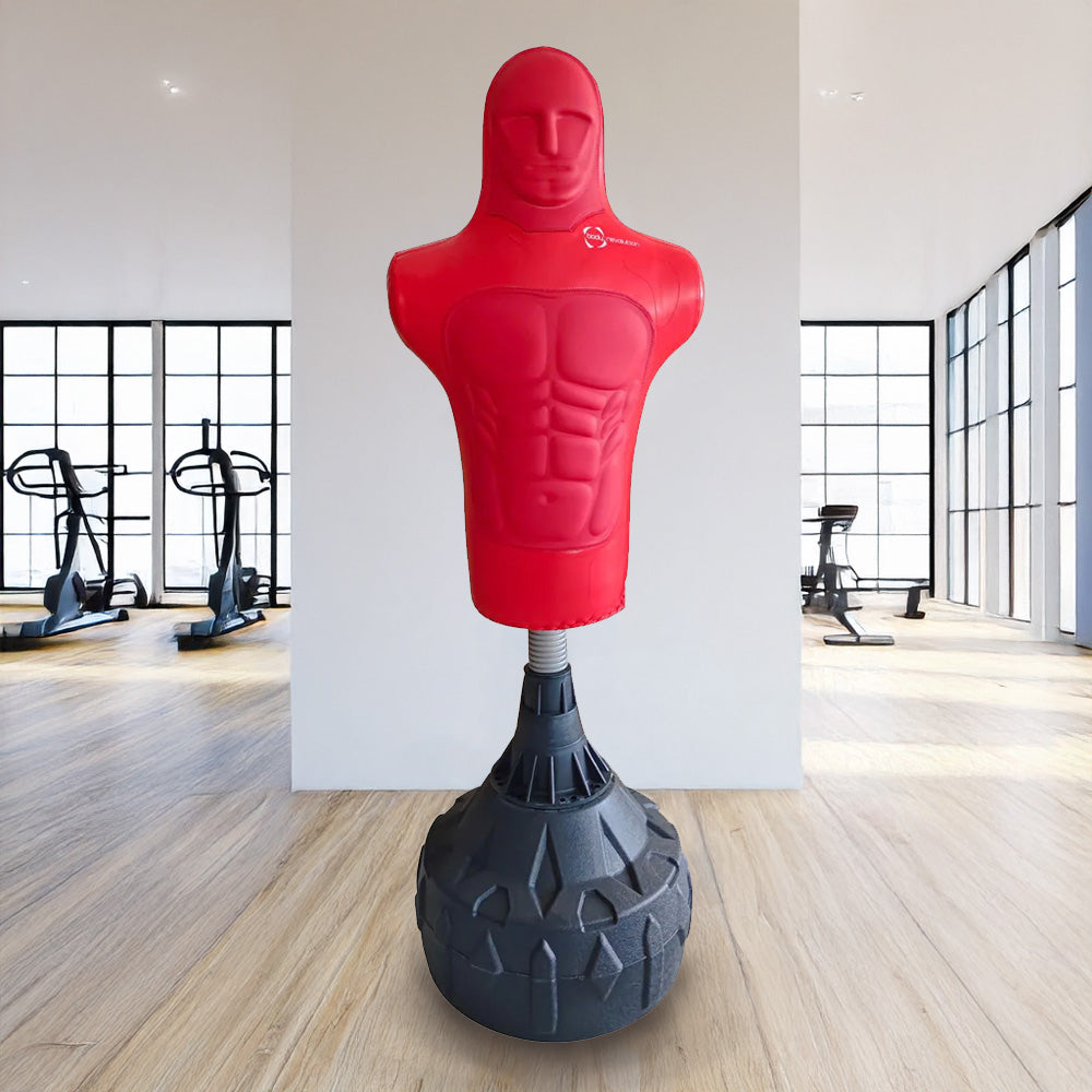 Un saco de boxeo rojo con forma humana está en un gimnasio con equipo al fondo.