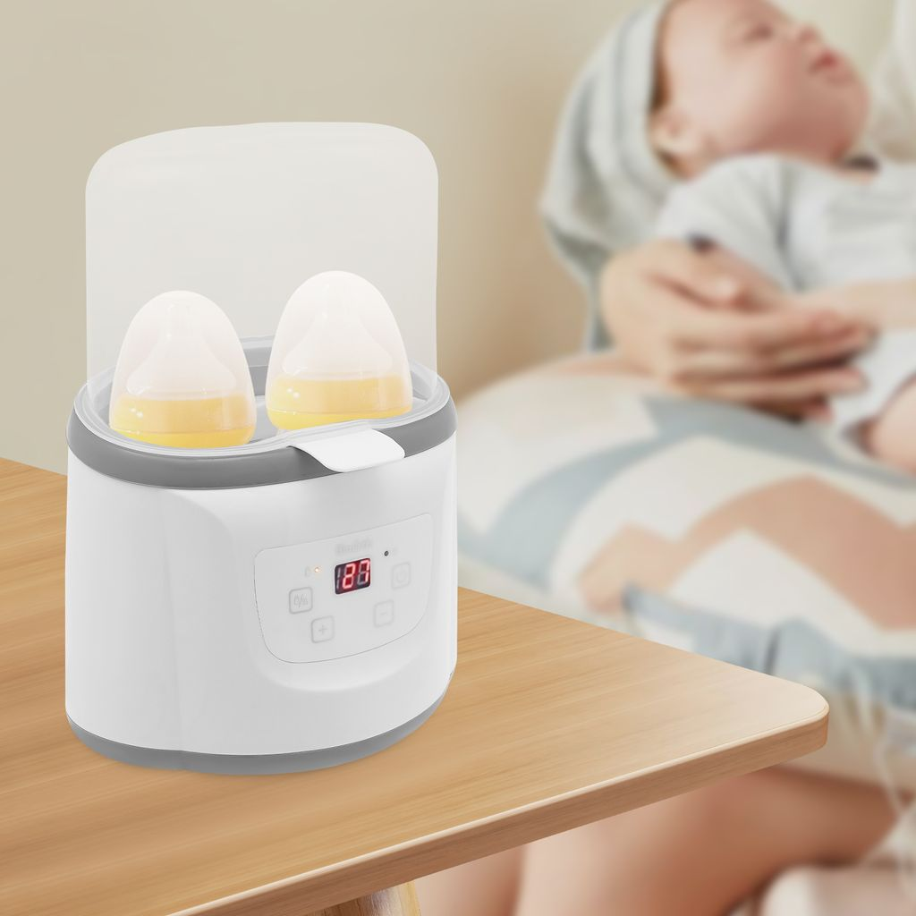 Ein weißer Flaschenwärmer mit zwei Flaschen darin, ein Baby im Hintergrund.