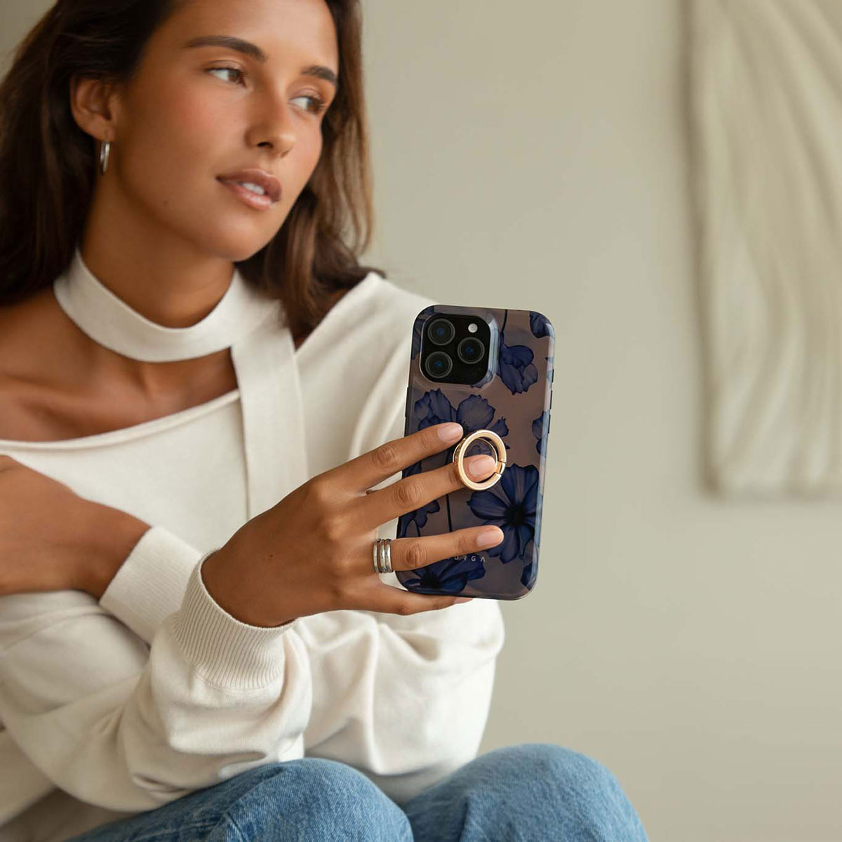 Una mujer con suéter blanco sostiene un teléfono con diseño floral azul y un anillo dorado.