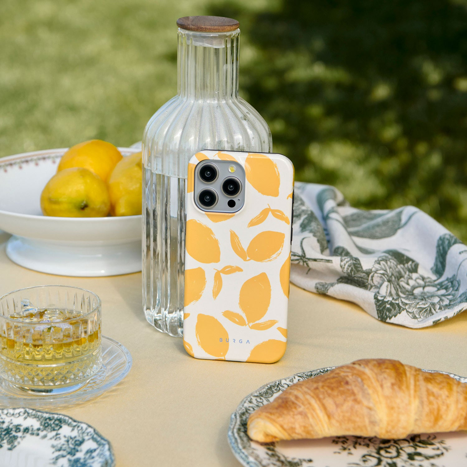 A phone with lemon pattern sits on a table with a croissant, glass of tea, and lemons.