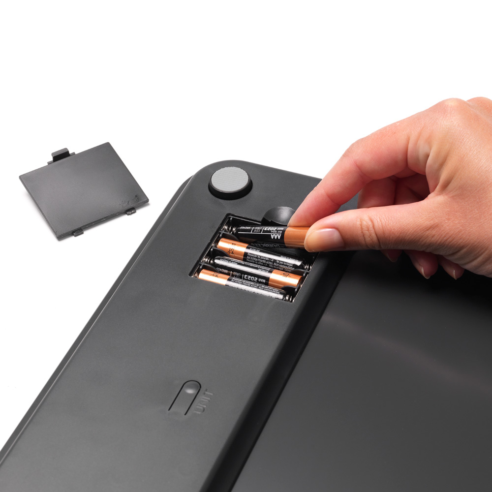Una mano inserisce una batteria AAA in una bilancia. Il coperchio è accanto.