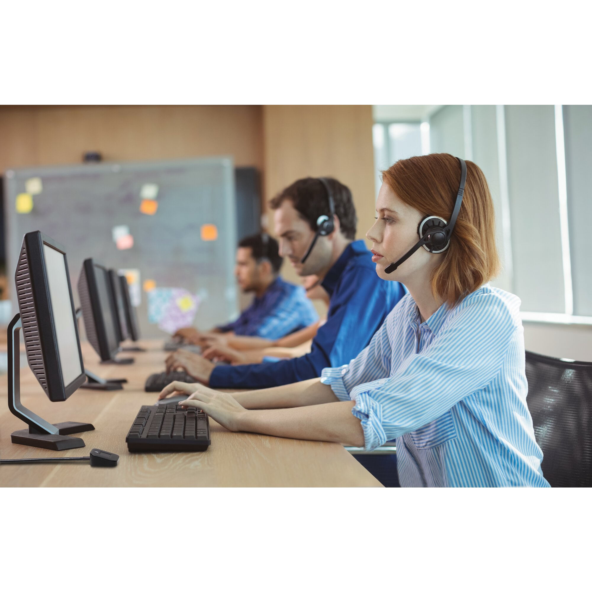 Menschen mit Headsets arbeiten nebeneinander an Computern. Fokus auf die Frau vorne.
