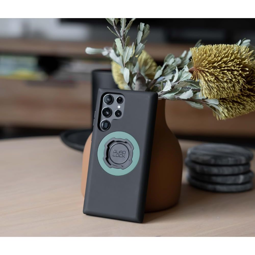 Uno smartphone nero con supporto Quad Lock vicino a un vaso di fiori secchi.