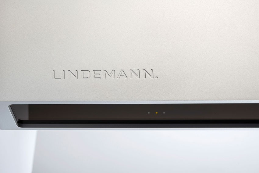 Wytłoczone logo Lindemann na szczotkowanej powierzchni metalowej z ciemnym pasem i wskaźnikami poniżej.