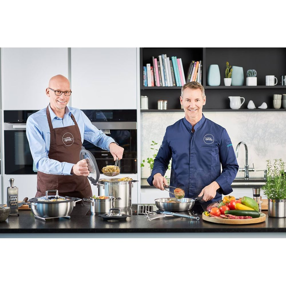 Zwei Männer kochen in einer modernen Küche. Einer hält ein Sieb mit Artischocken, der andere brät etwas in einer Pfanne.