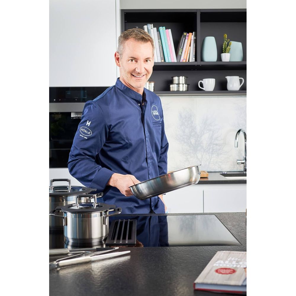 Ein Koch in blauer Uniform hält eine Bratpfanne in einer modernen Küche mit Kochgeschirr.
