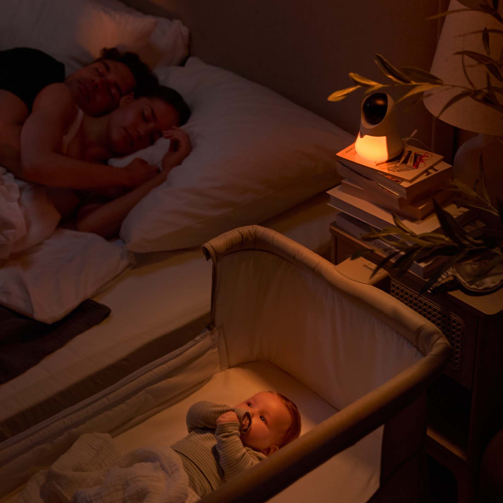 Ein Baby in einem Beistellbett, Eltern schlafen im Hintergrund. Ein Babyphone leuchtet.
