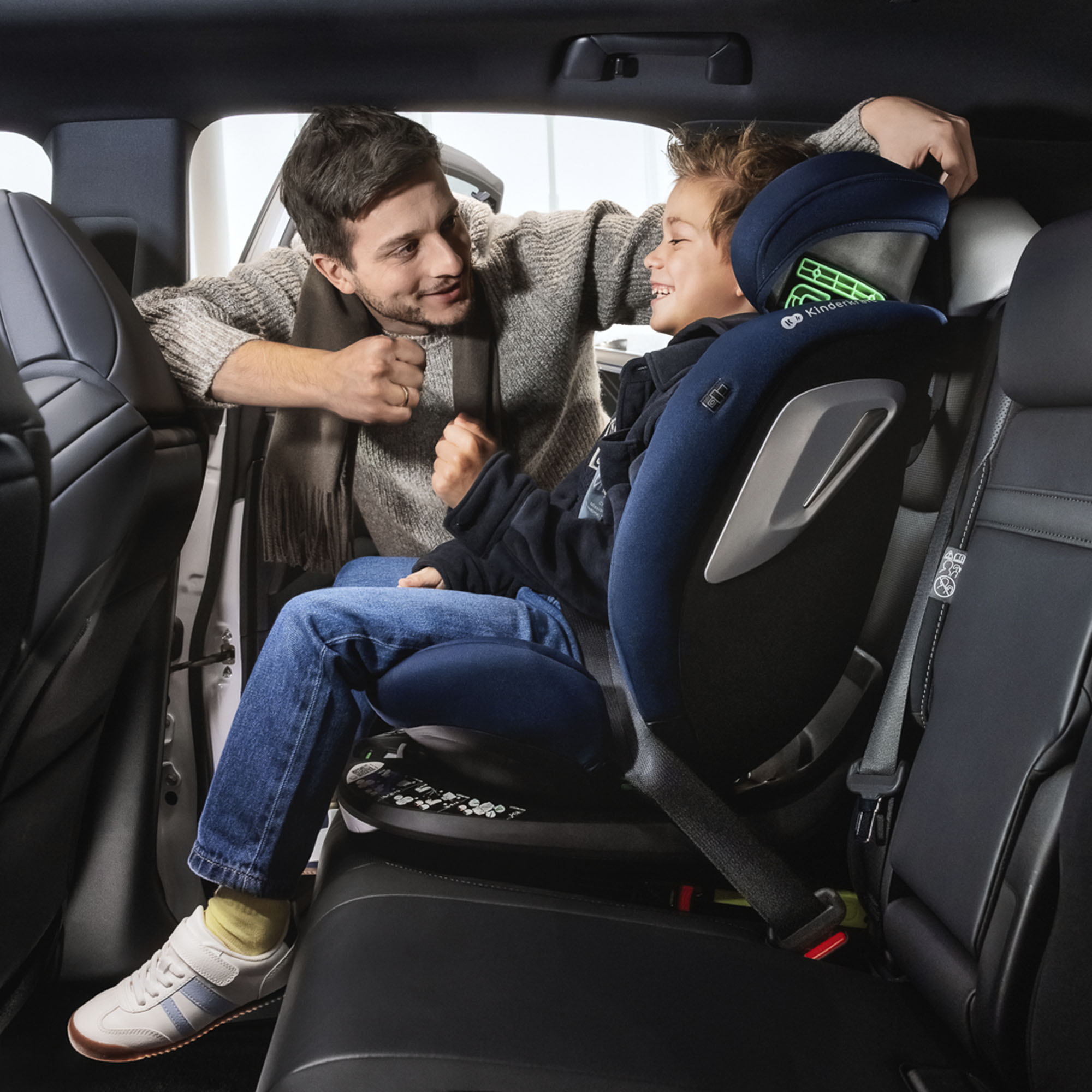 Hombre se inclina hacia niño en silla de auto, sonriendo.