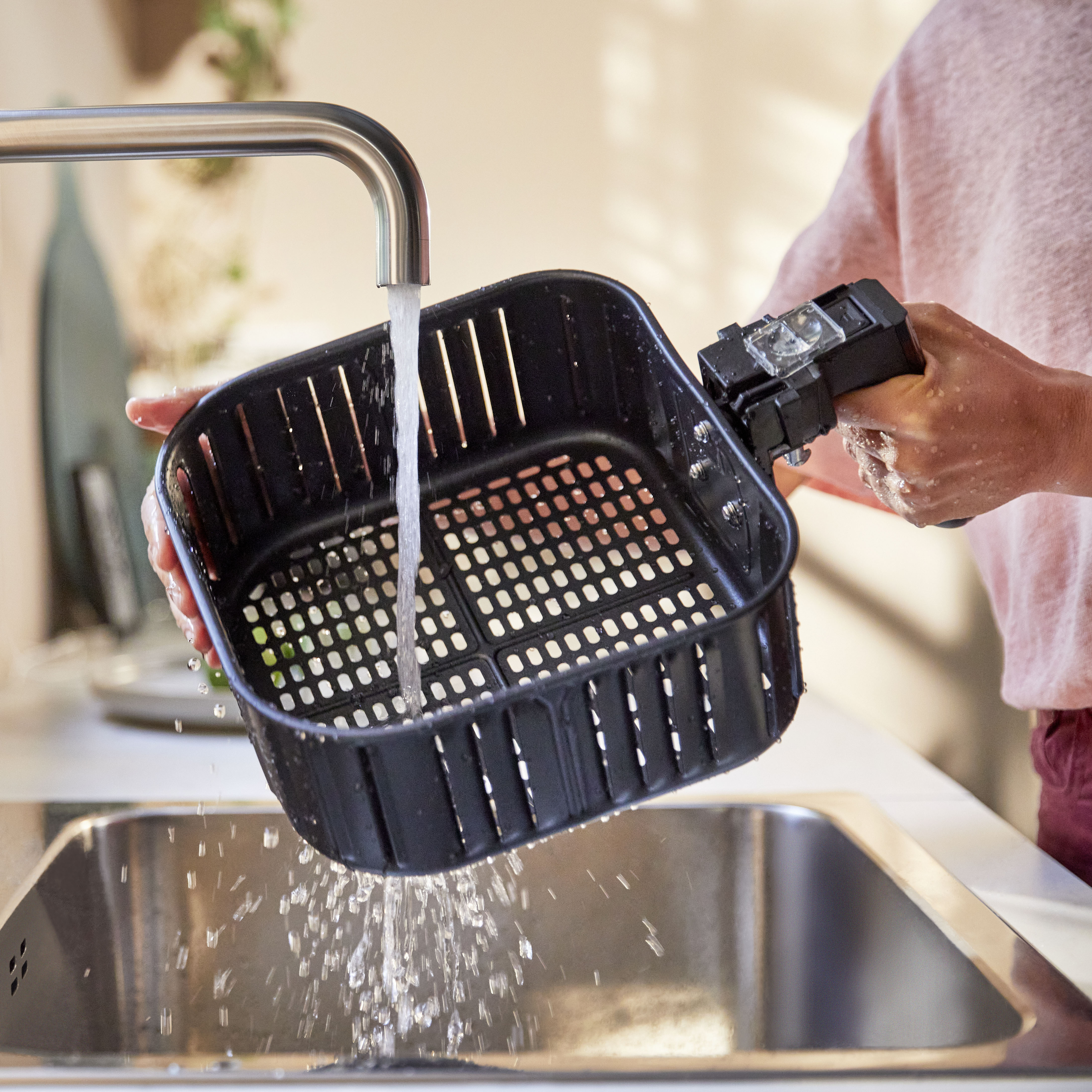 A person rinses a black air fryer basket under running water in a sink.