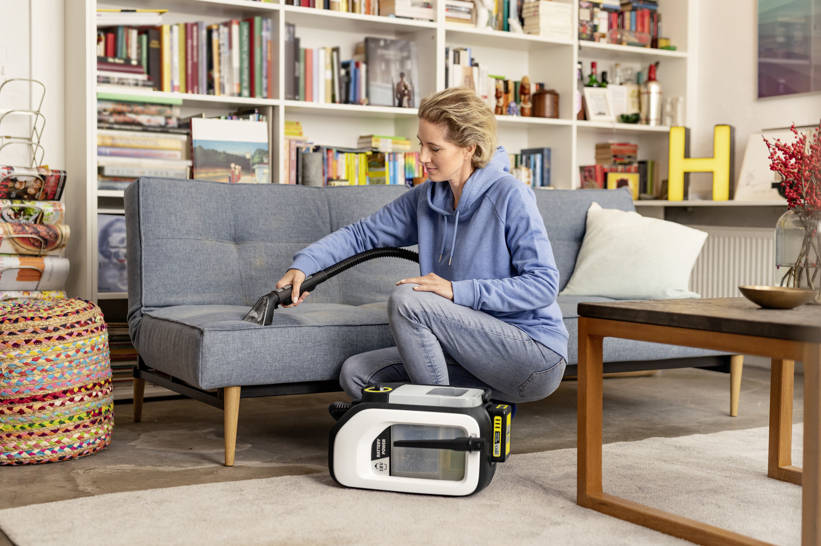 Donna pulisce divano con aspiratore a spruzzo davanti a libreria con pouf colorato e tavolino.
