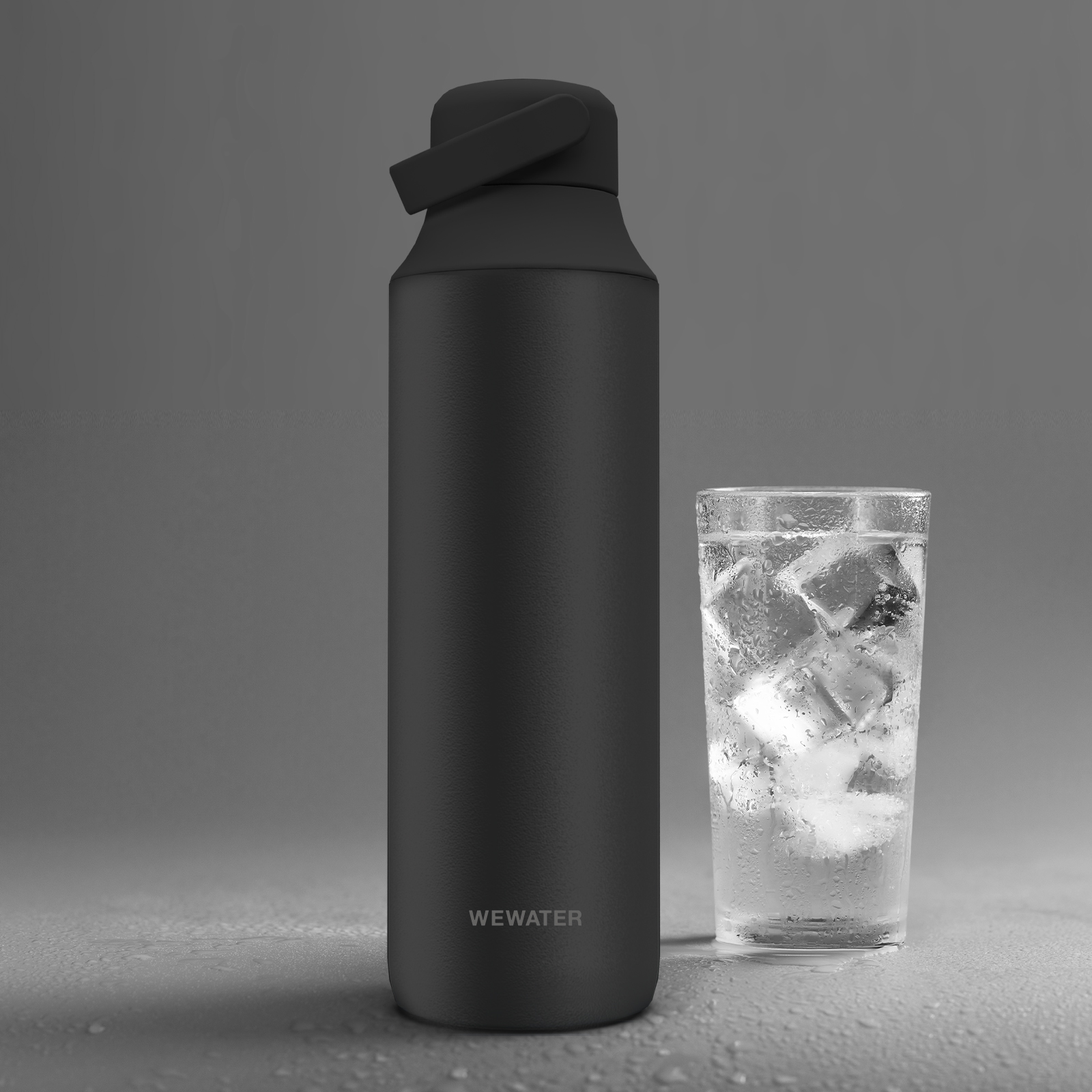 Botella de agua negra junto a un vaso con agua helada sobre fondo gris.