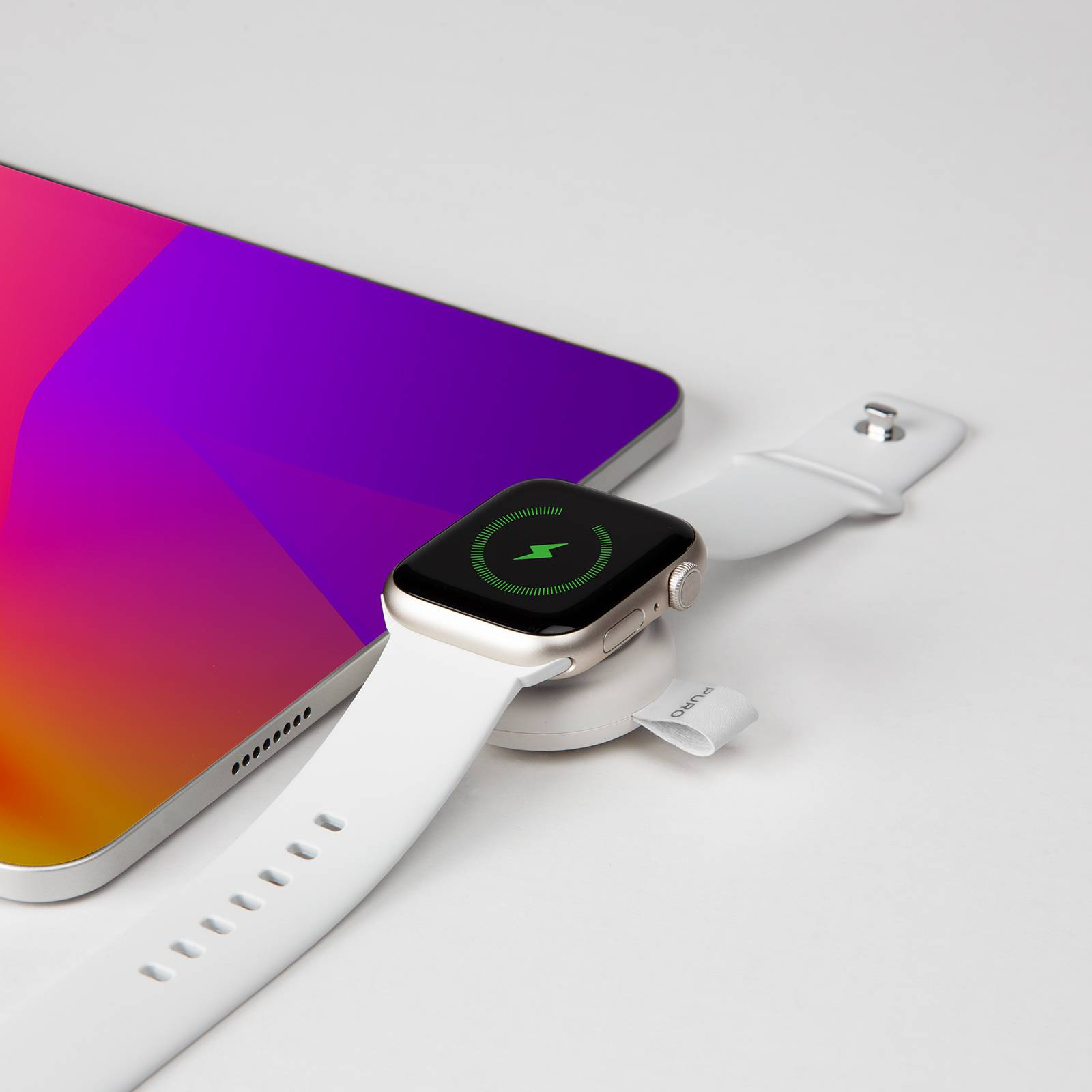 Un Apple Watch plateado sobre un cargador junto a una tablet con pantalla morada/naranja.