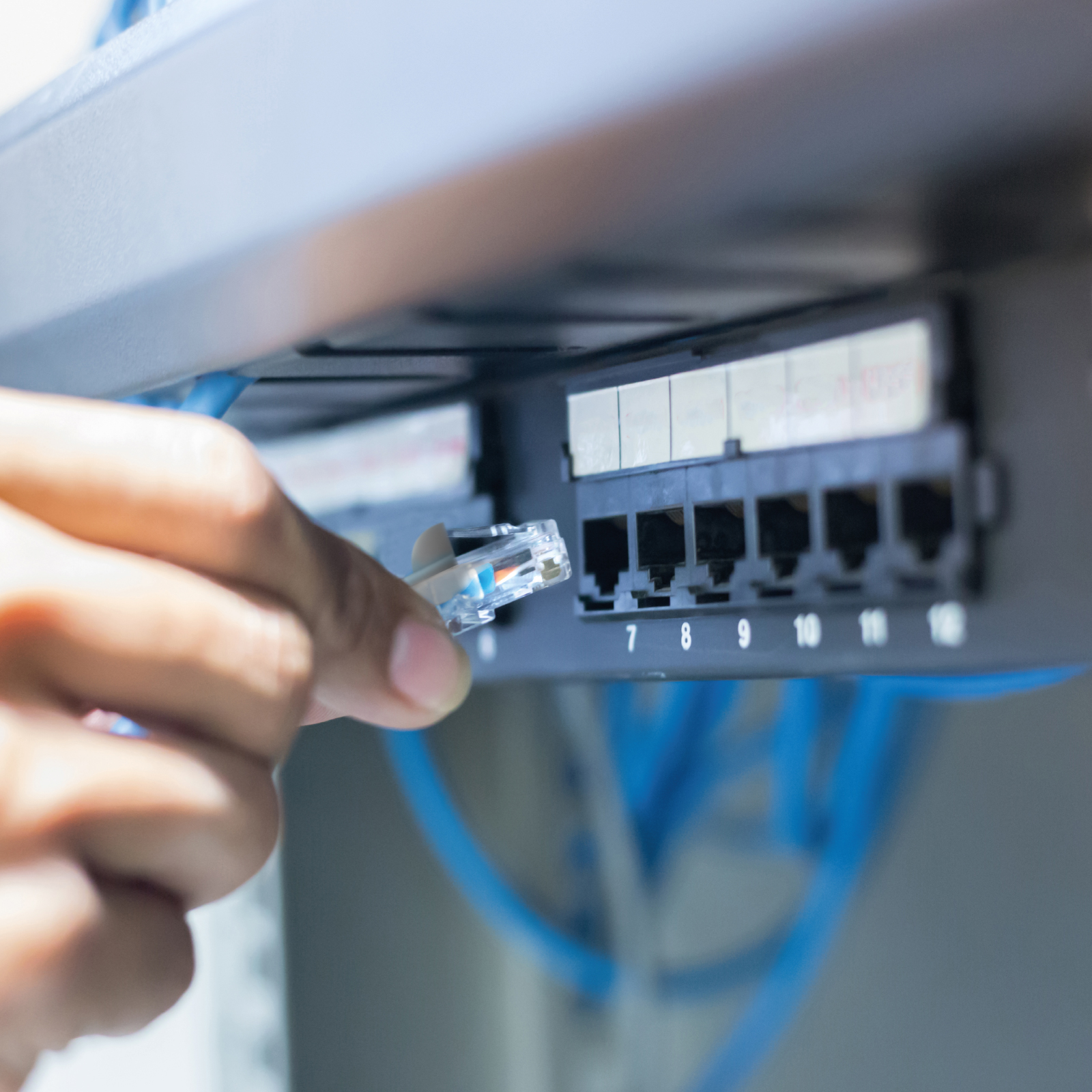 Una mano sujeta un conector de cable de red cerca de un rack de red con cables azules.