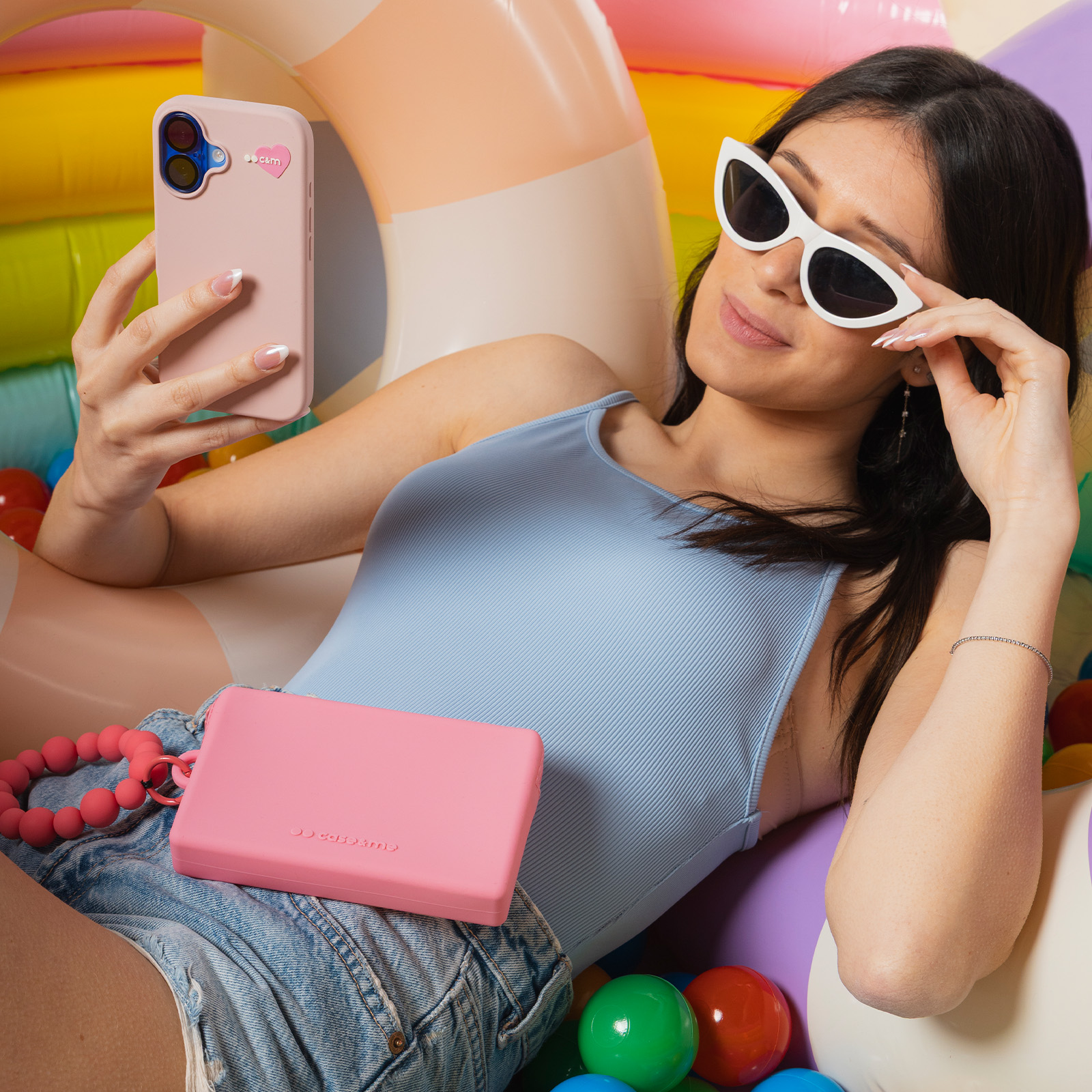 Mujer recostada con teléfono, traje de baño azul y gafas de sol blancas, rodeada de pelotas.