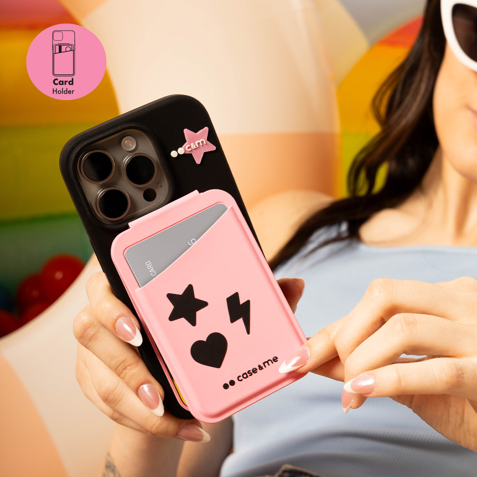 Una mano sostiene un smartphone con funda negra y tarjetero rosa con pegatinas de estrella y corazón.