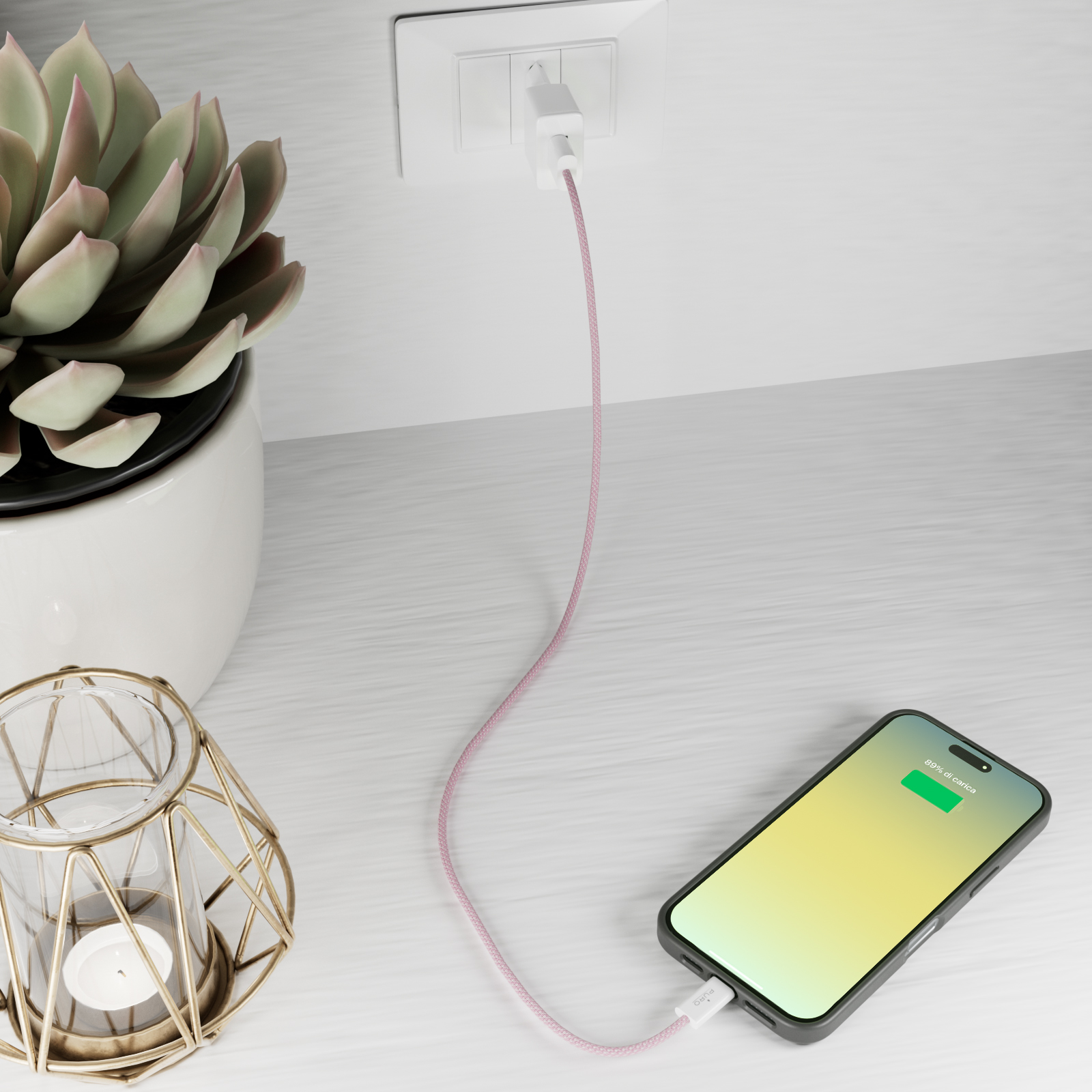 Un telefono si carica da una presa con cavo rosa. Una pianta e una candela sono vicine.