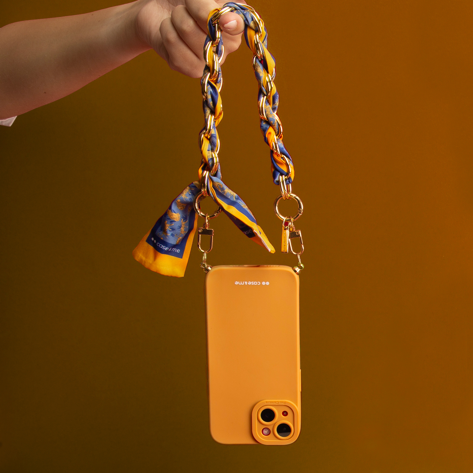 Una mano tiene una cover arancione per telefono con catena dorata e foulard.