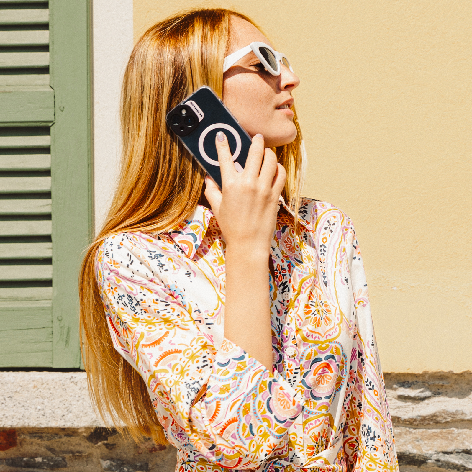 Una mujer con cabello rubio largo y gafas de sol blancas sostiene un teléfono en su oído, vistiendo un vestido estampado colorido.