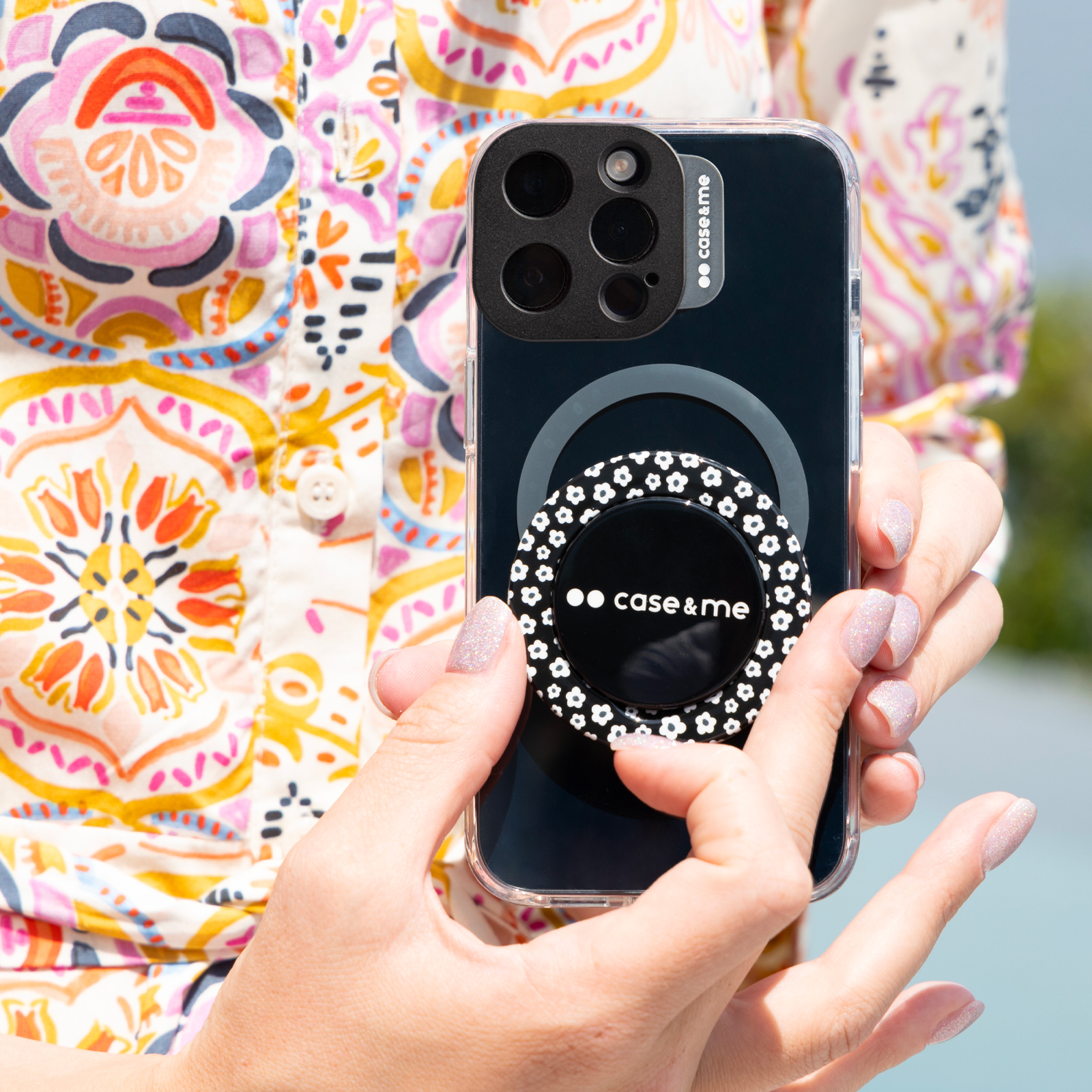 Manos sostienen un teléfono negro con módulo de cámara y agarre floral ante camisa colorida.