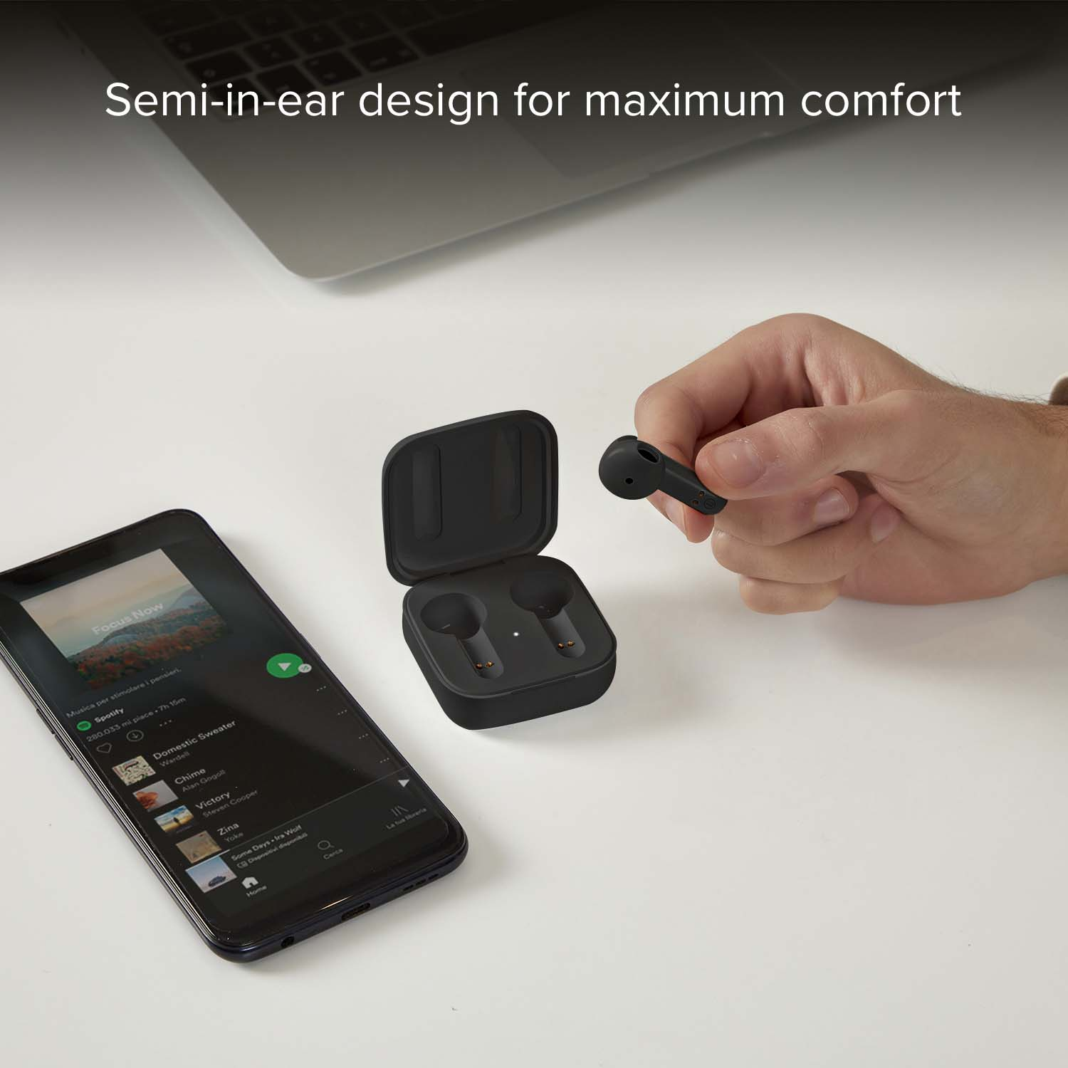 Una mano sostiene un auricular negro sobre un estuche de carga abierto junto a un smartphone.