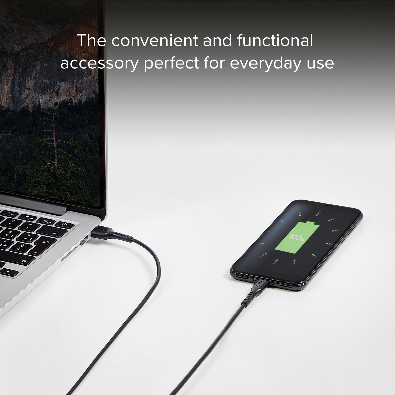 A laptop charges a smartphone displaying 100% battery.