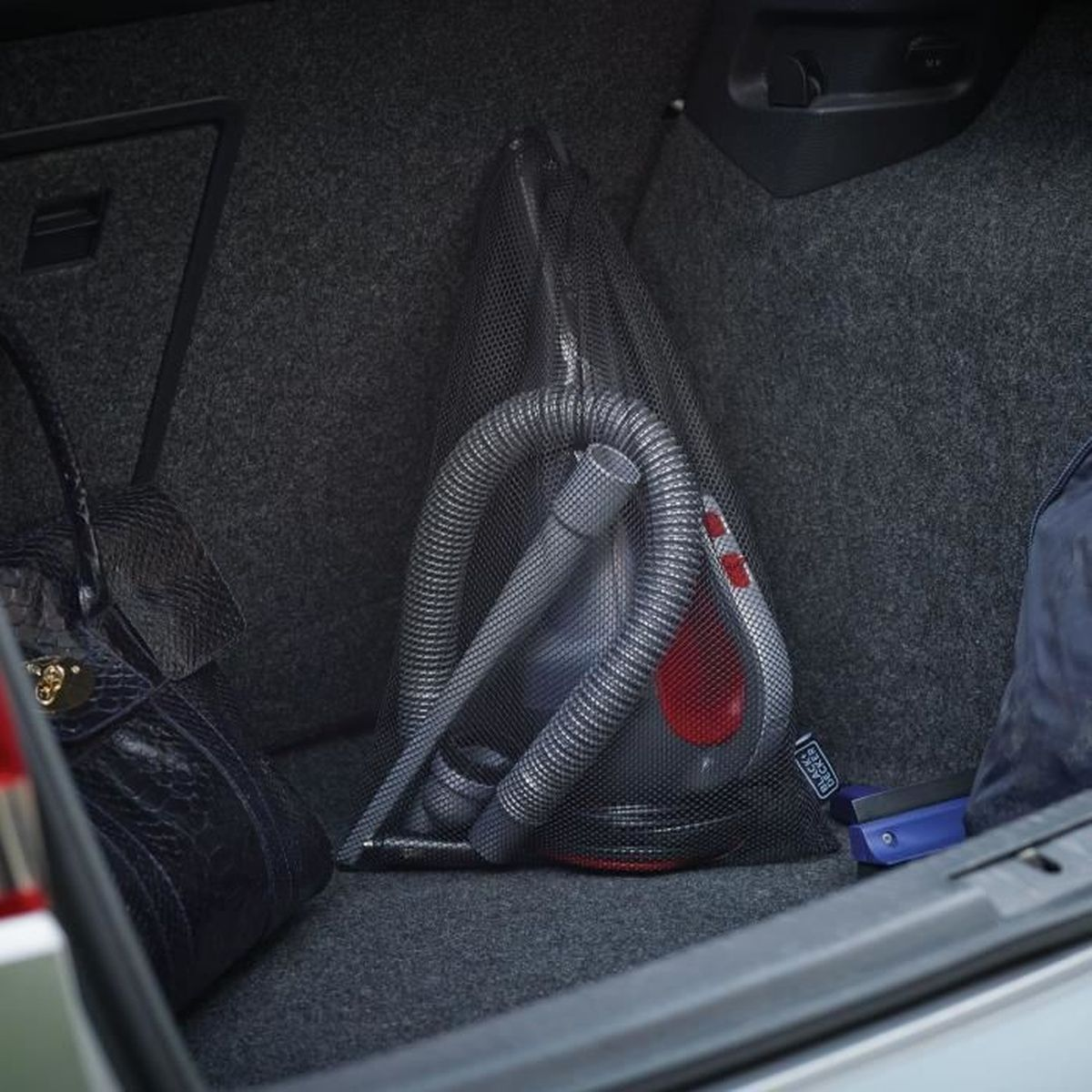 Ein schwarz-roter Autostaubsauger mit grauem Schlauch, in einem Netzbeutel verstaut, liegt im Kofferraum eines Autos.