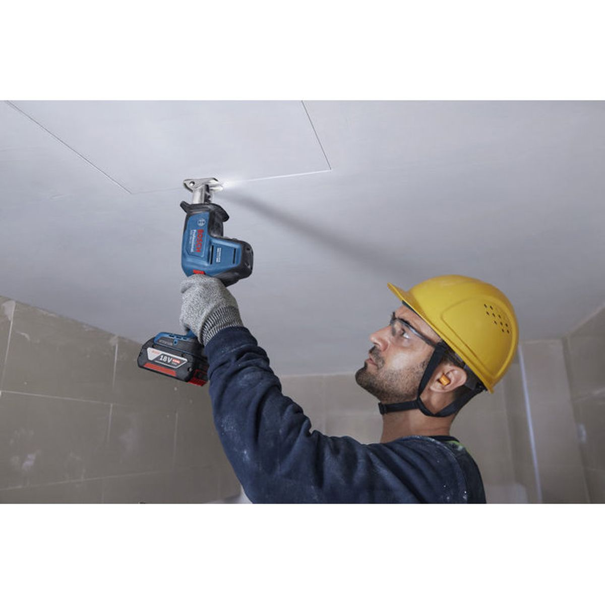 Ein Bauarbeiter mit gelbem Helm schneidet mit einer blauen Säbelsäge eine quadratische Öffnung in eine weiße Decke.